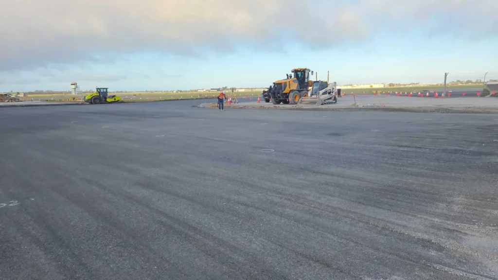 A wide view of a construction site with heavy machinery, including a roller and a grader, and a worker in a safety vest standing on freshly paved asphalt under a partly cloudy sky.