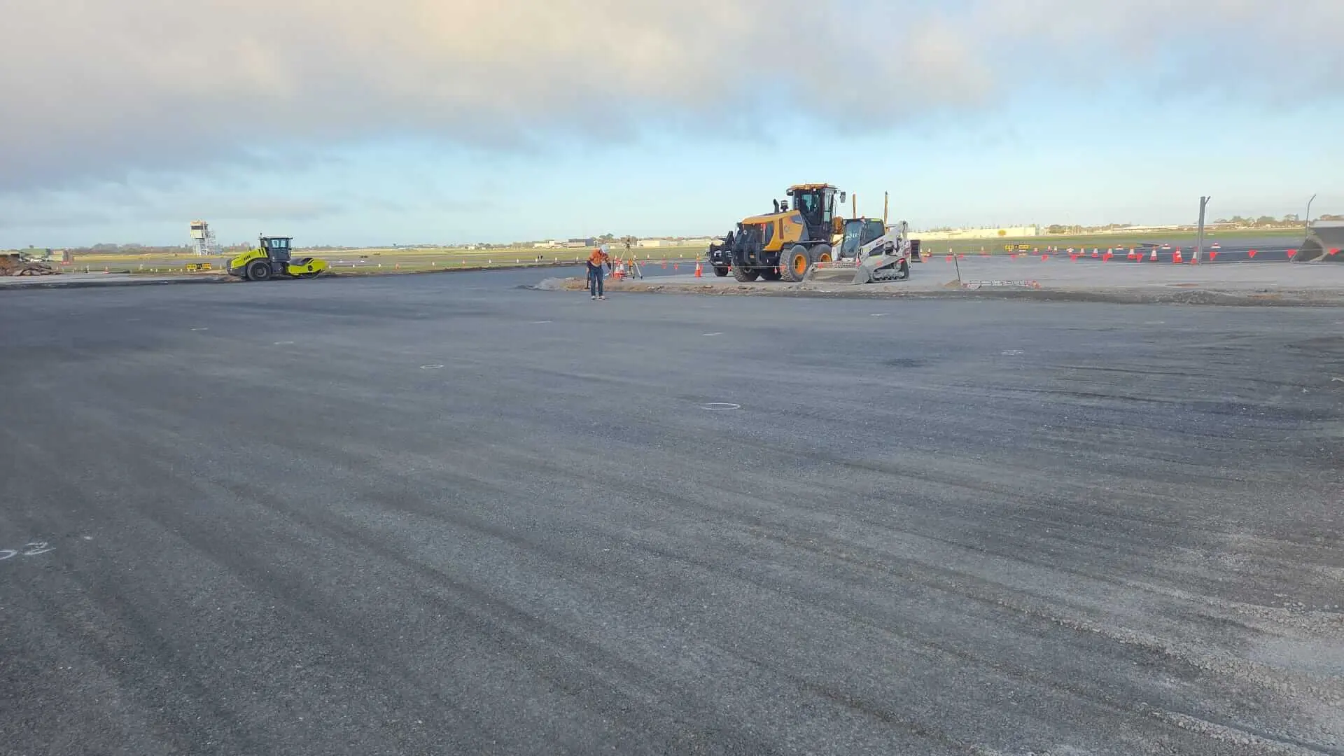 A wide view of a construction site with heavy machinery, including a roller and a grader, and a worker in a safety vest standing on freshly paved asphalt under a partly cloudy sky.