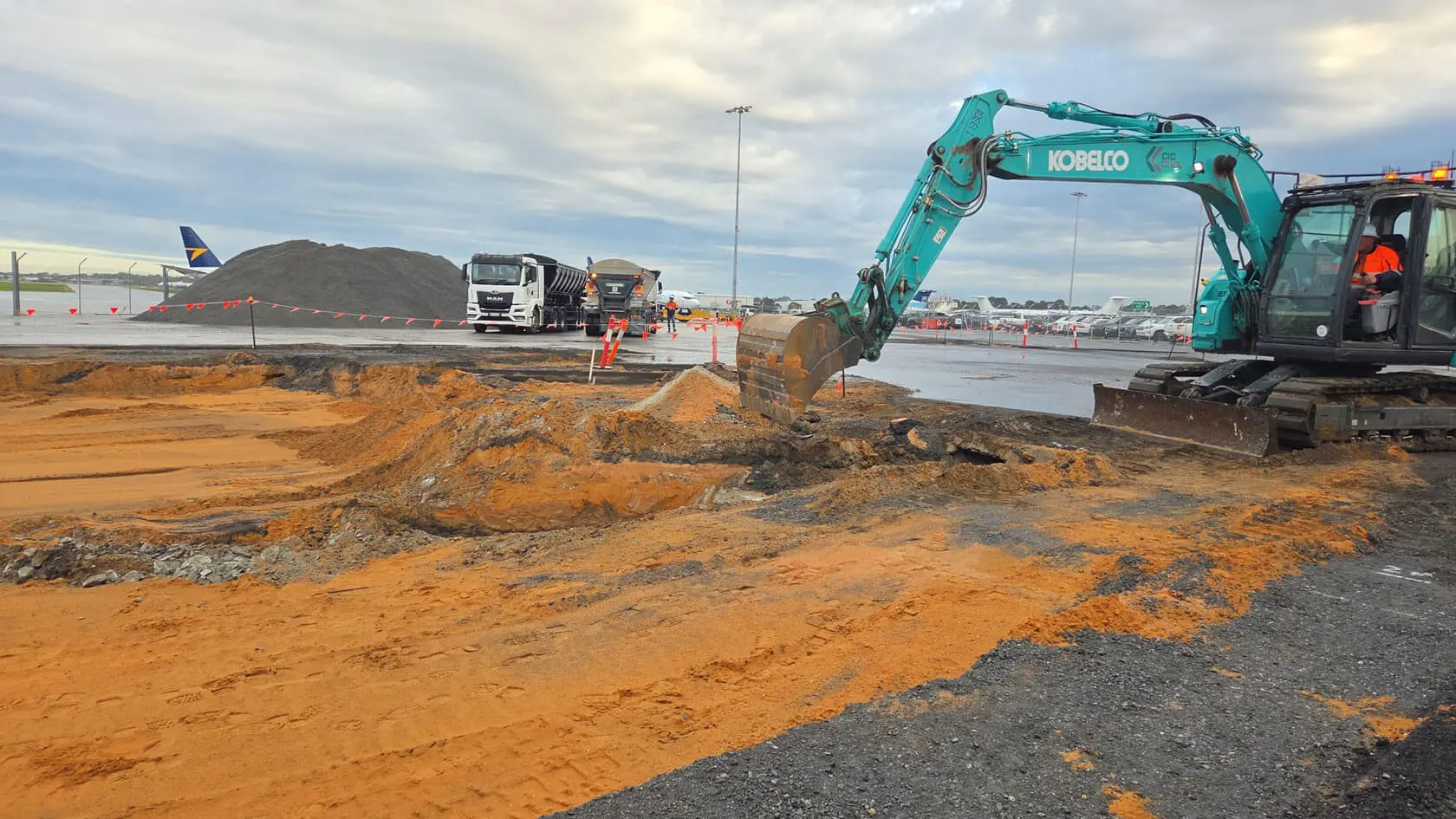 A blue excavator digs earth at a construction site near an airport runway, with trucks and piles of dirt in the background and orange safety barriers along the work area.