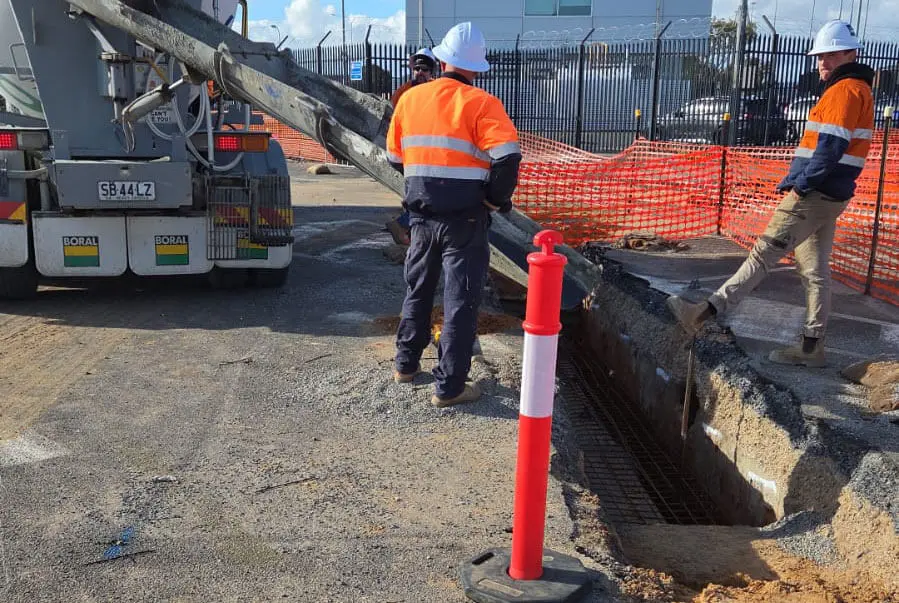 Two construction workers in safety gear guide wet concrete from a cement truck chute into a trench at a fenced construction site. One worker stands on the edge of the trench while the other manages the chute.
