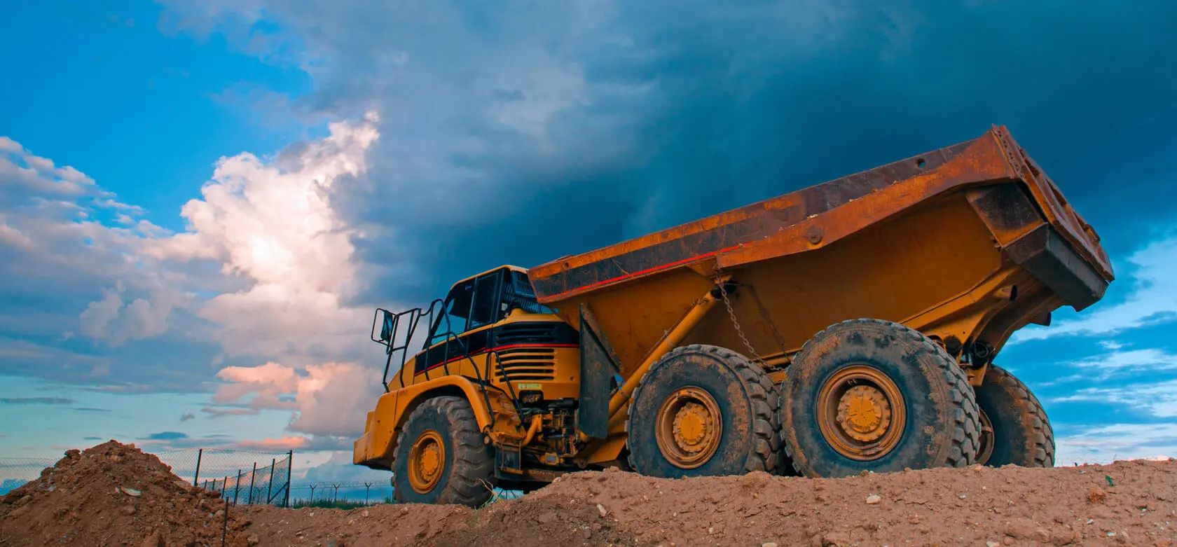 A large yellow dump truck sits on a dirt mound under a dramatic, cloudy sky, with sunlight illuminating part of the clouds in the background.