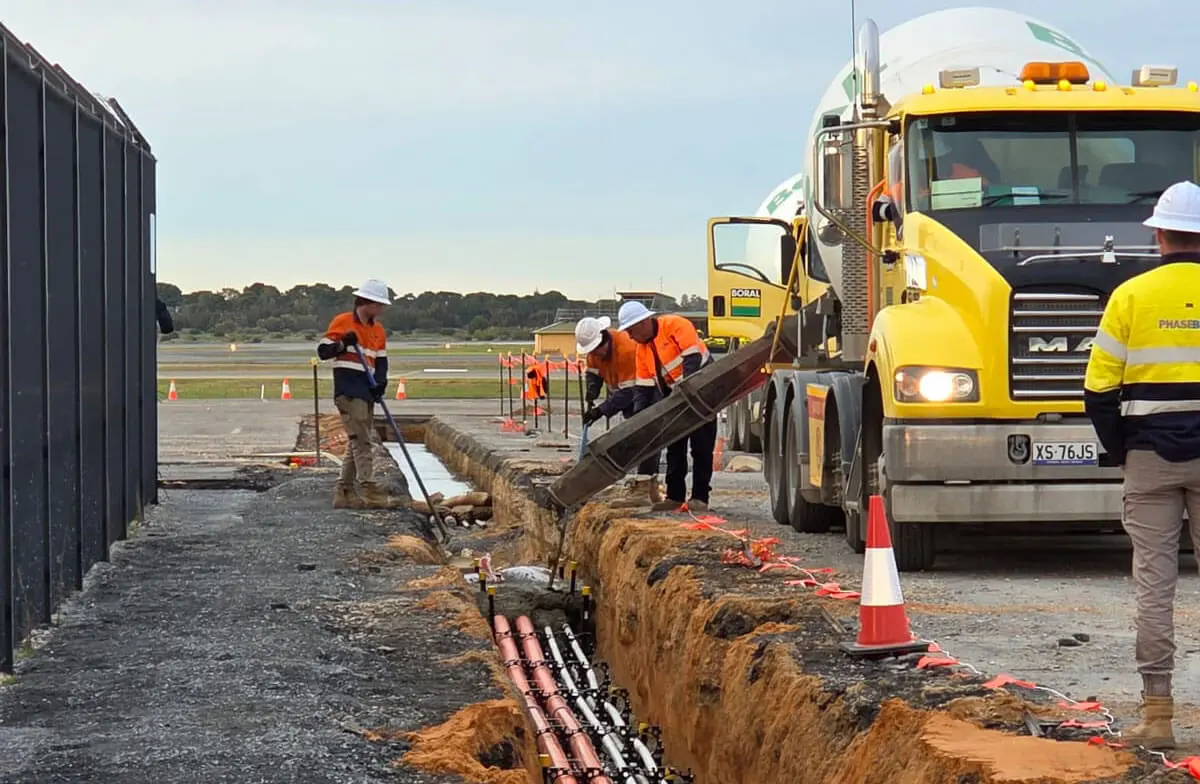 Several construction workers in safety gear pour concrete from a yellow cement truck into a trench containing cables at a worksite, with traffic cones and fencing nearby.