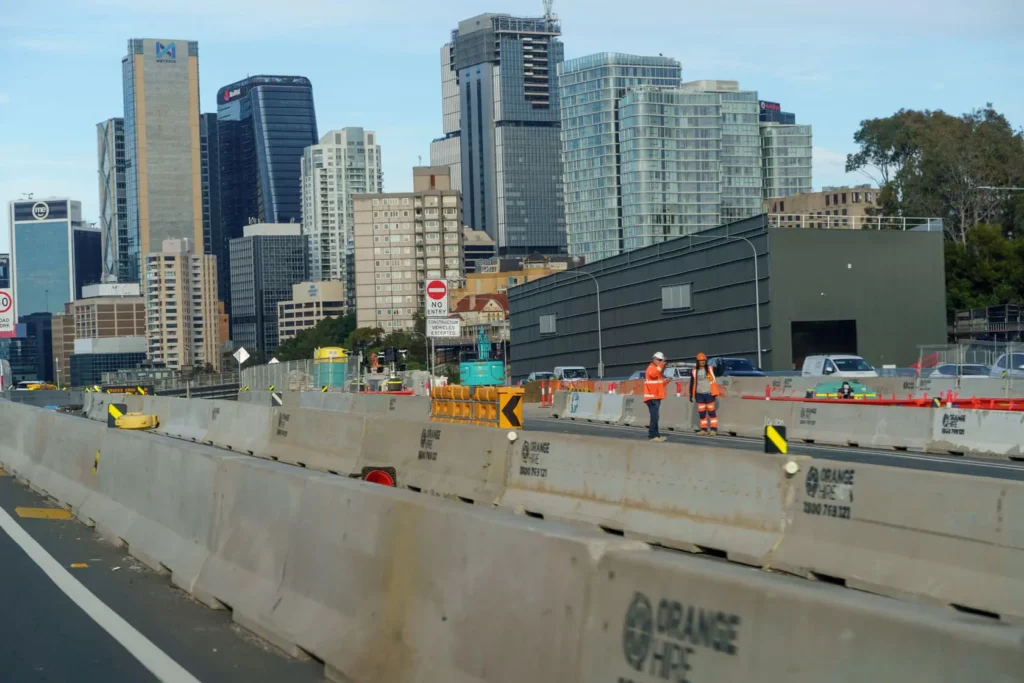 Construction workers in safety gear work on a city street closed off by concrete barriers, with tall modern buildings and skyscrapers in the background.