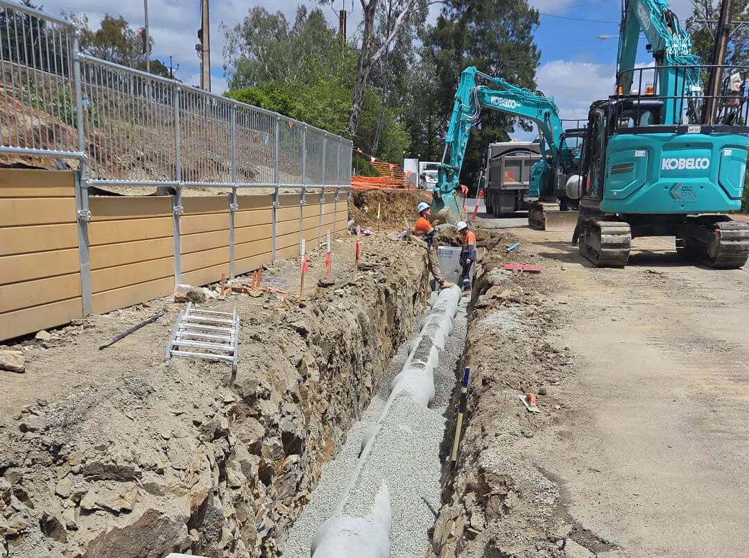 Construction workers install pipes in a deep trench next to a fenced retaining wall. Excavators and construction equipment are nearby on a dusty worksite under a partly cloudy sky.
