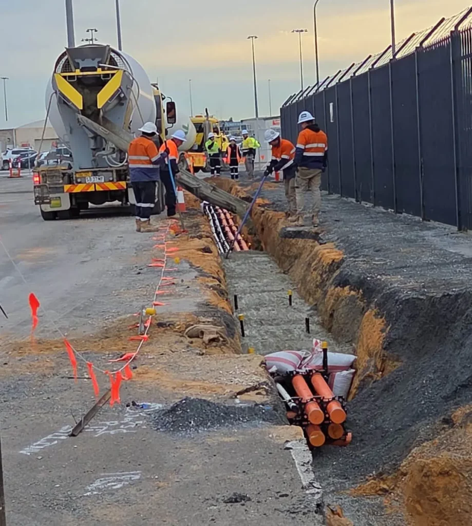 Workers in high-visibility clothing install orange utility pipes in a long trench beside a fenced road. A concrete mixer truck is on site, and safety barriers with red flags line the work area.