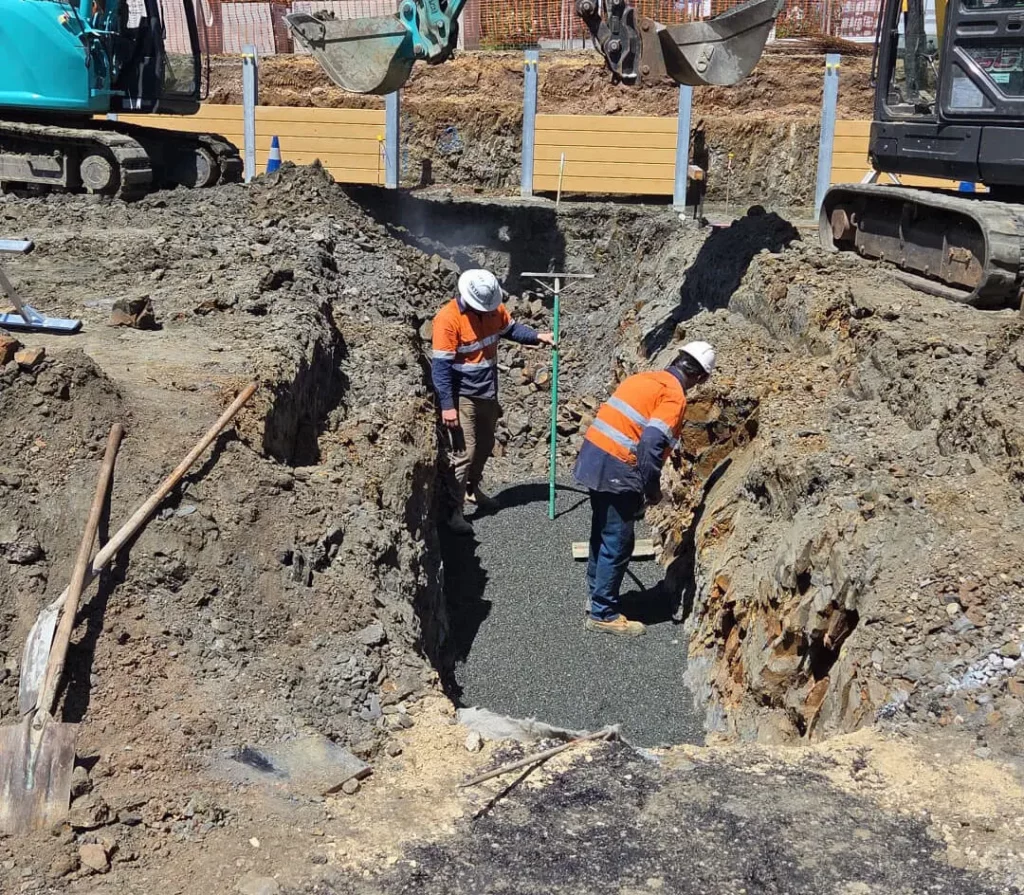 Two construction workers wearing hard hats and orange safety vests work in a deep trench, shoveling gravel as machinery and tools surround them on an active construction site.
