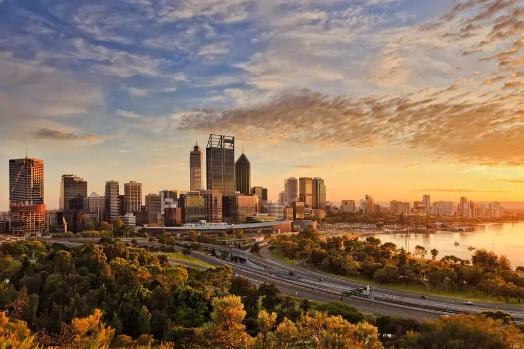 A city skyline with tall buildings and skyscrapers at sunset, with a river, bridges, and lush green trees in the foreground under a partly cloudy sky.