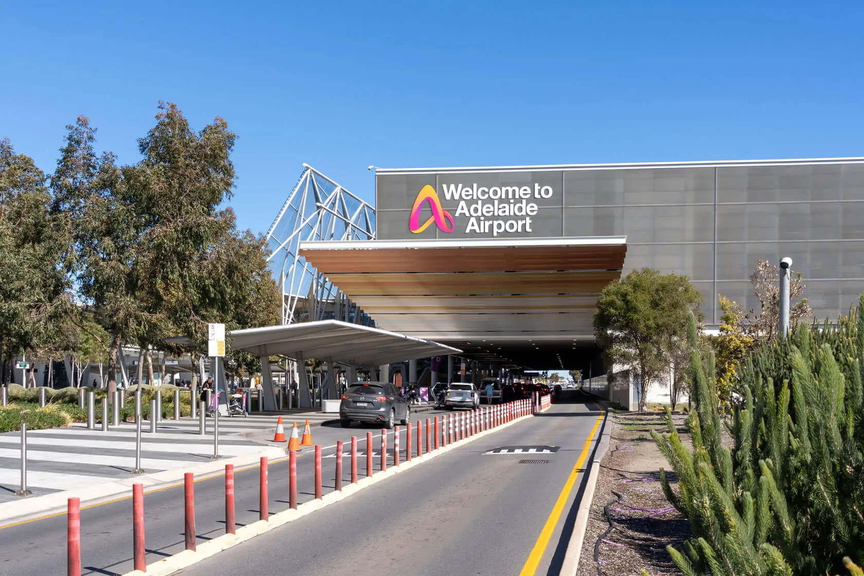 The entrance of Adelaide Airport with cars dropping off passengers under a covered area, trees along the road, and a large sign reading "Welcome to Adelaide Airport" on the building.