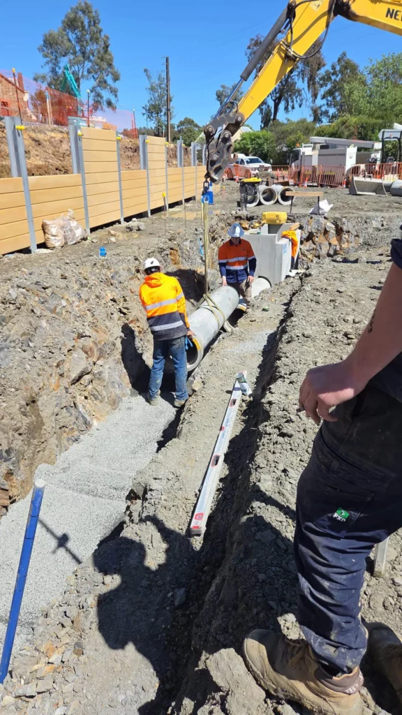 Four construction workers in safety gear are installing large concrete pipes in a gravel trench, with an excavator assisting; tools and construction materials are visible around the site.