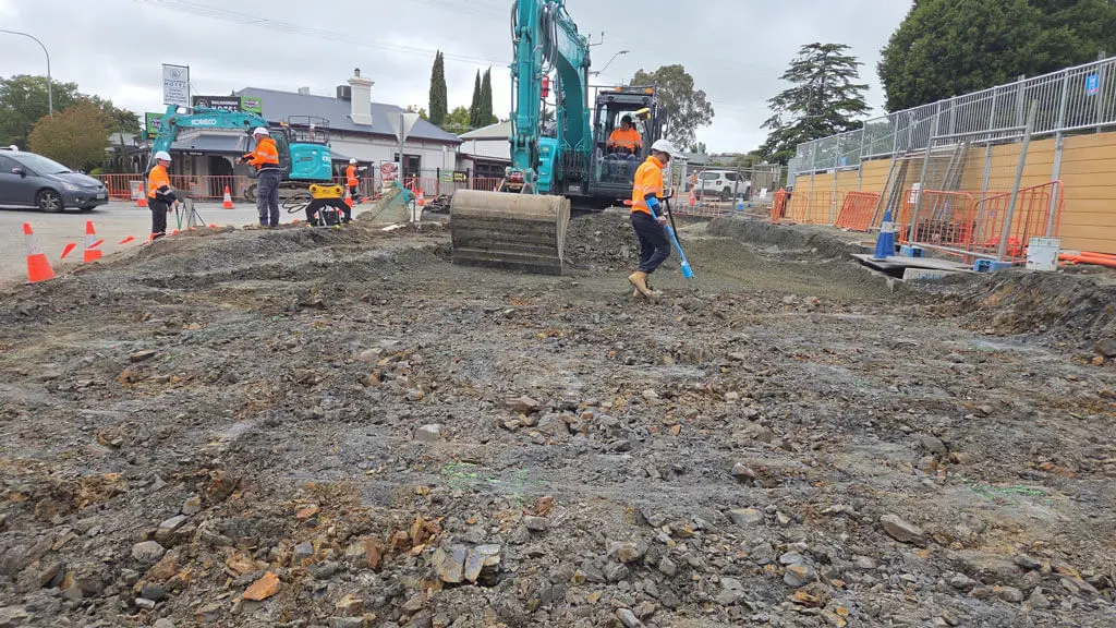 Construction workers in orange safety vests operate machinery and use tools on a rough, muddy construction site near a street, with traffic cones, fencing, and nearby buildings visible in the background.