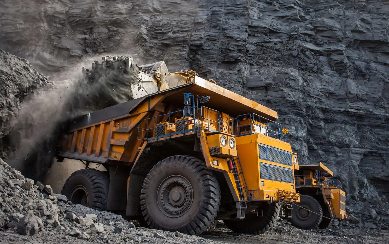 A large yellow mining dump truck is being loaded with gravel and rocks by heavy machinery at a quarry, with another dump truck in the background against a rocky cliff.