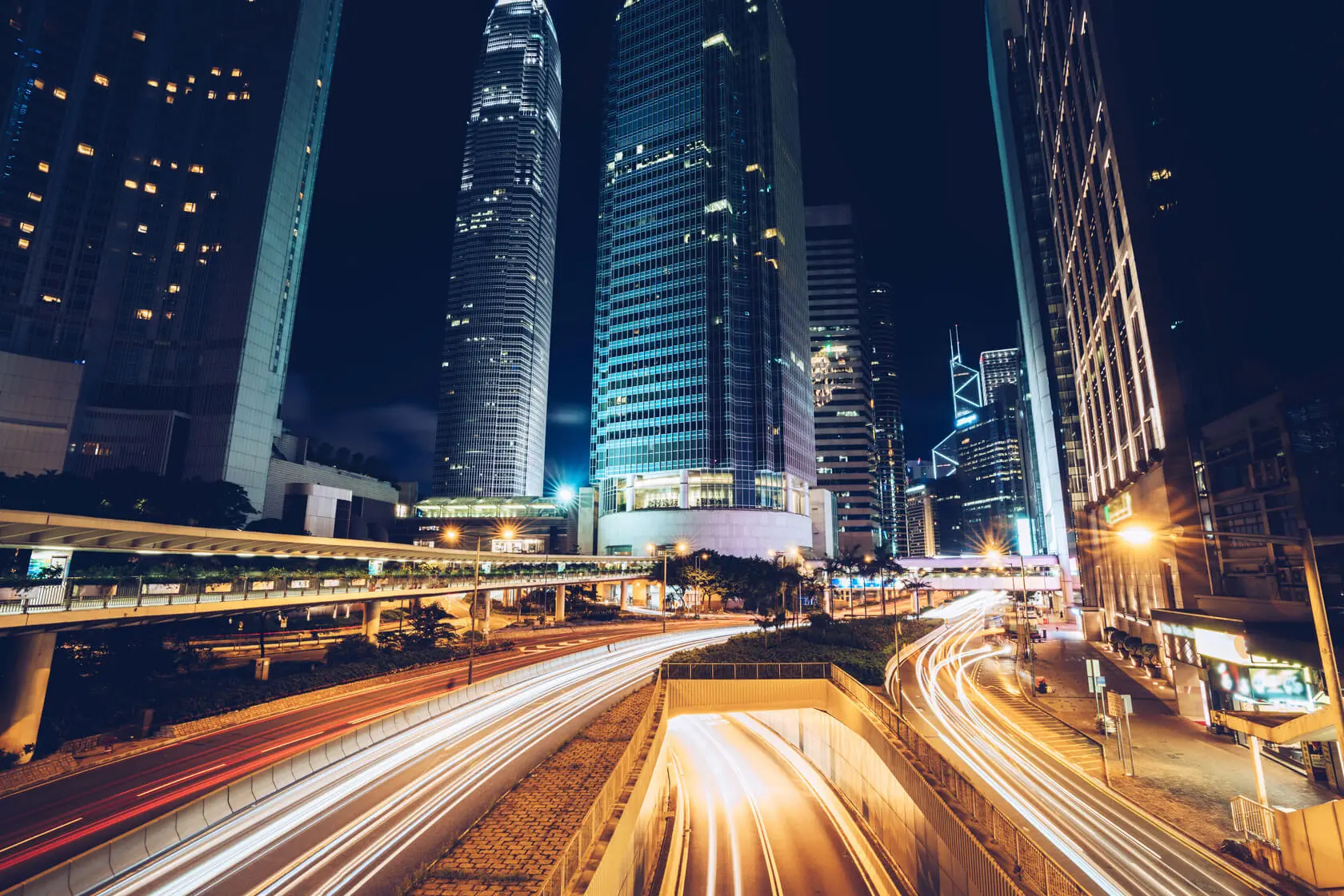 Nighttime cityscape with tall modern skyscrapers, illuminated lights, and blurred light trails from moving vehicles on winding roads, creating a dynamic sense of motion in an urban setting.