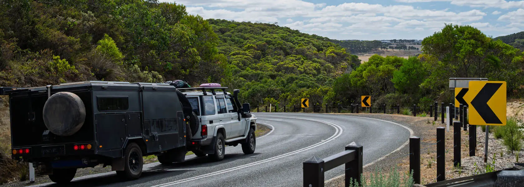 A white SUV towing a black trailer drives along a curving road bordered by trees and greenery. Multiple yellow road signs with black arrows indicate a sharp bend ahead under a partly cloudy sky.