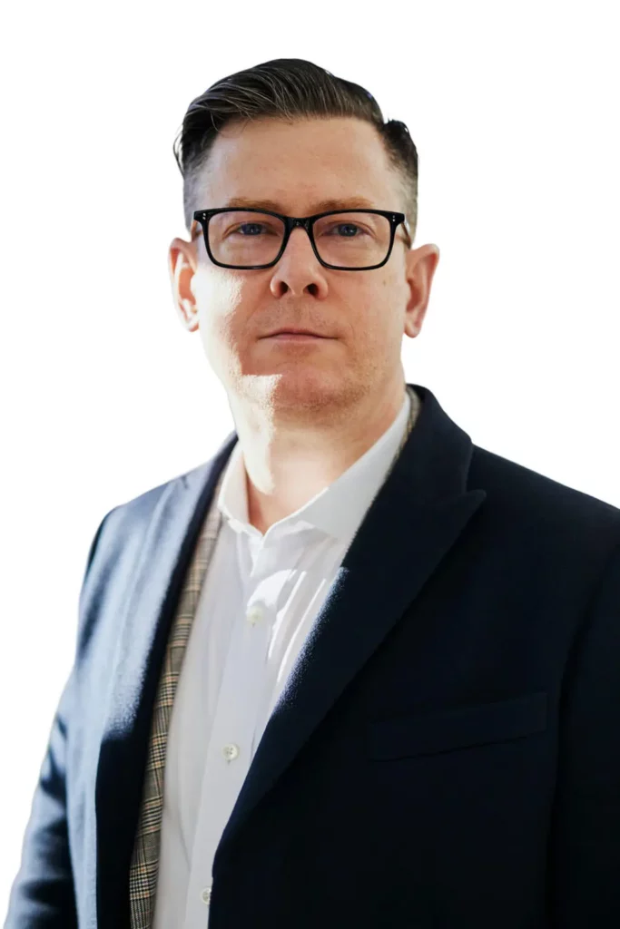 A man with short dark hair and glasses, wearing a dark blazer over a white shirt, stands facing the camera against a white background.