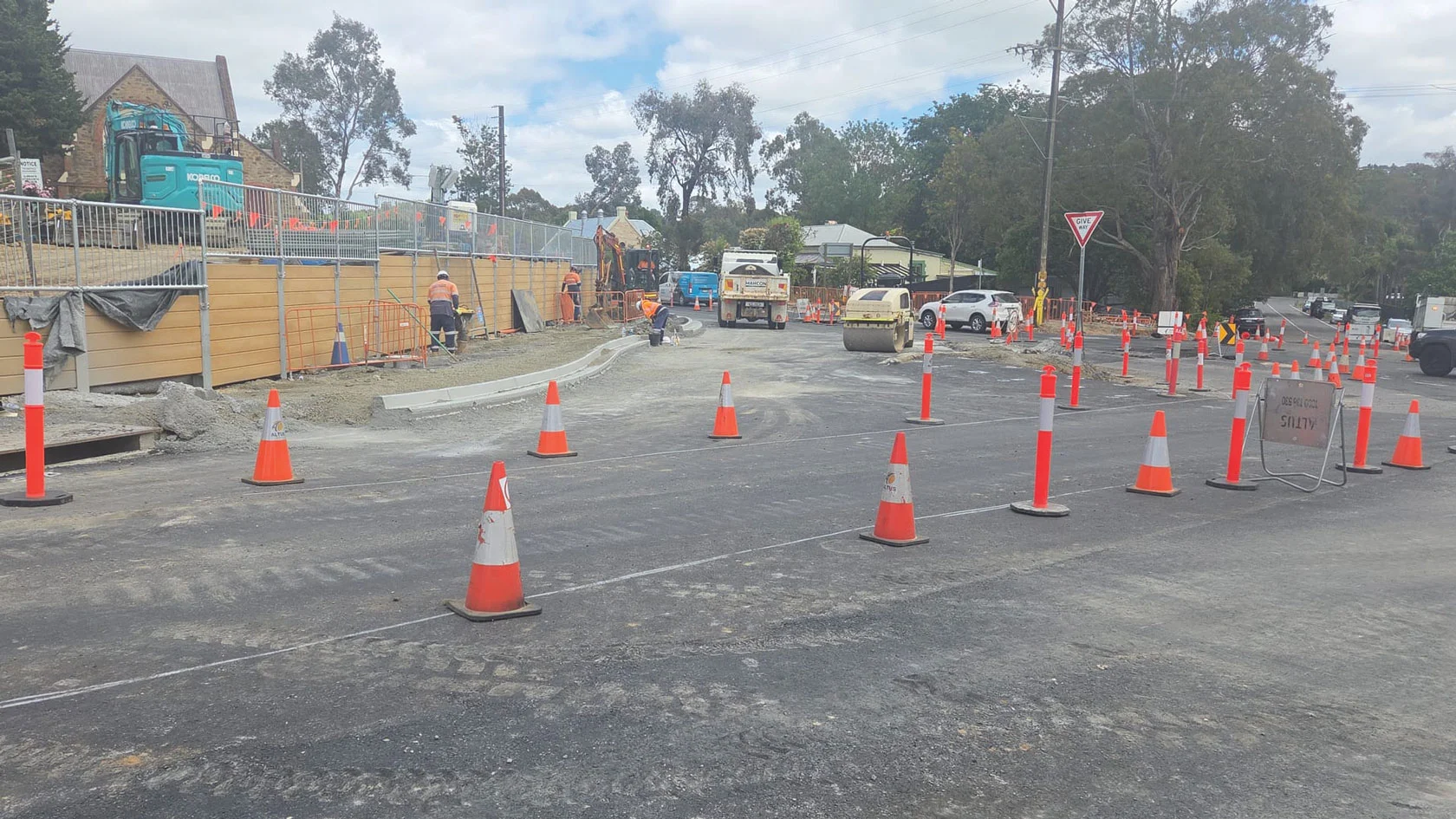 Road construction zone with orange cones, workers in safety gear, construction vehicles, and fencing. Fresh concrete and equipment are visible, along with a "Give Way" sign and cloudy sky in the background.